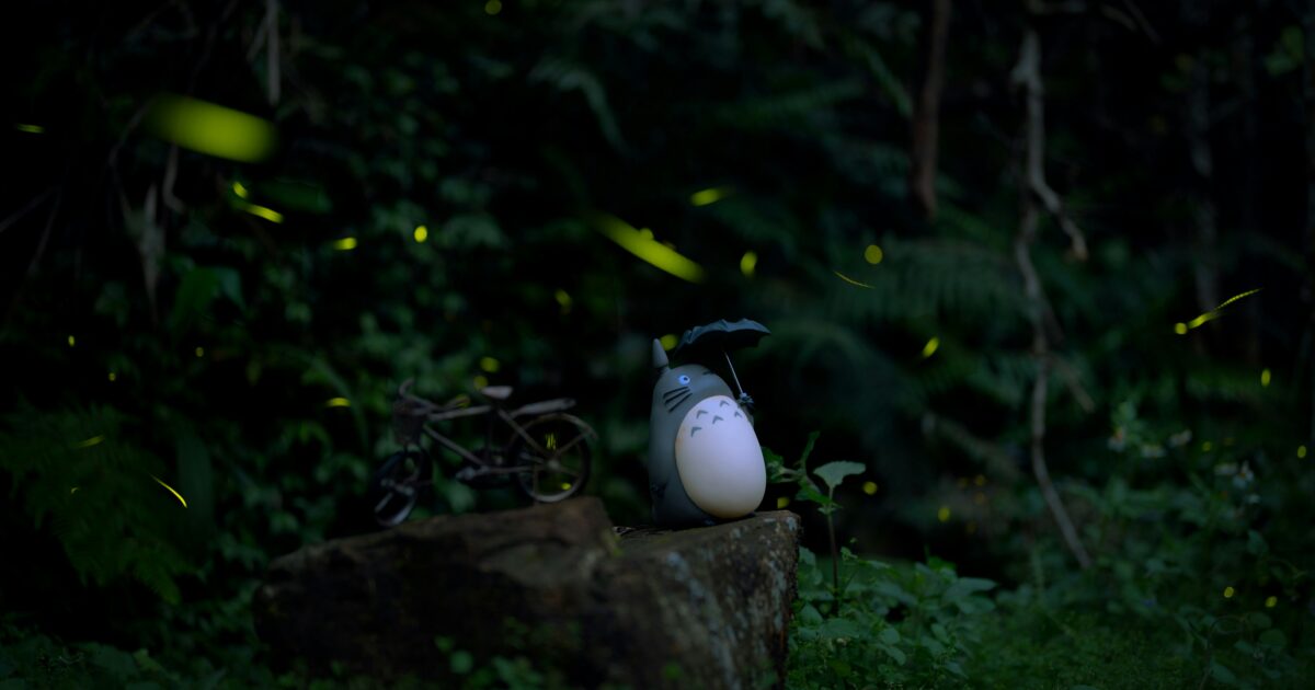 暗い森の中に置かれたトトロのフィギュアと自転車と蛍の光